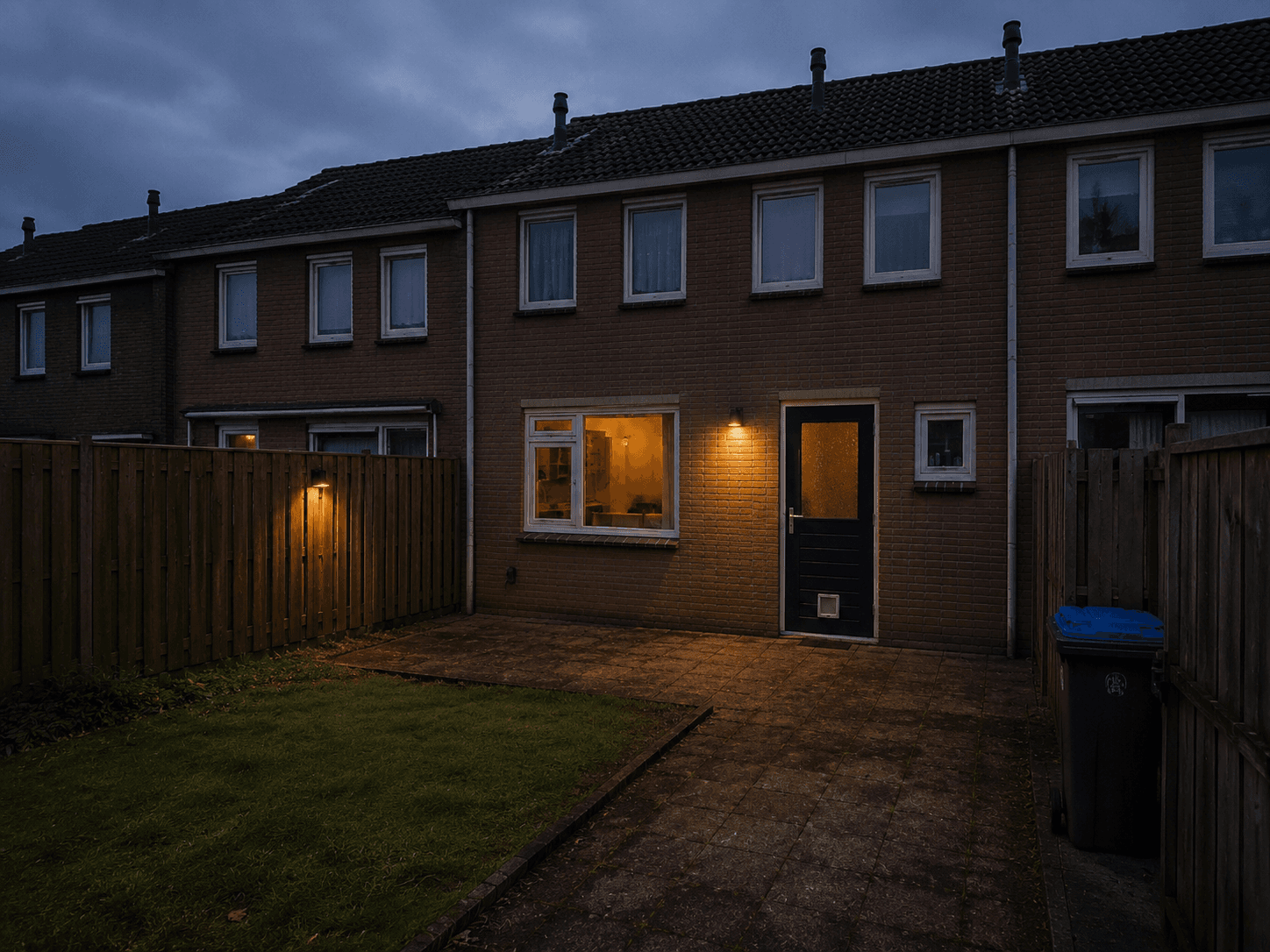 Originele achtertuin vóór de renovatie, met grijze tegels, grasstrook, houten schutting en buitenverlichting bij schemer.