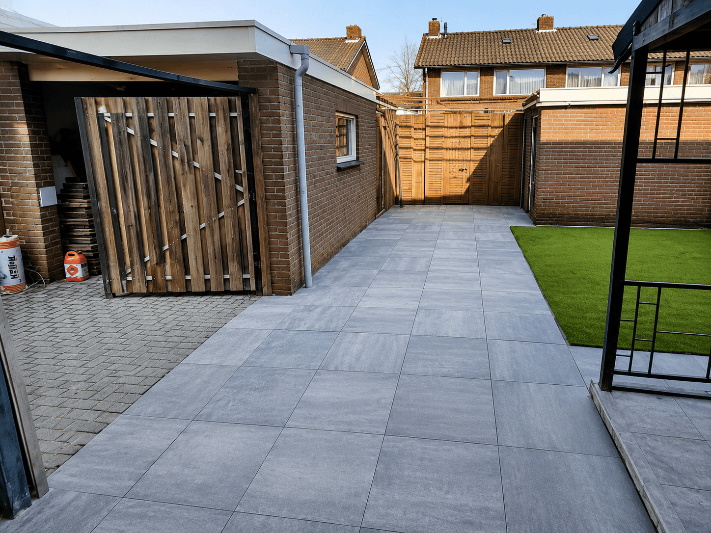 Achtertuin na de renovatie, met grijze keramische bestrating, kunstgras en een tuinprieel.