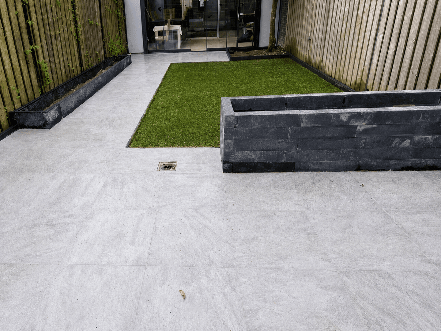Smalle stadstuin na de renovatie, met lichte bestrating, kunstgrasstrook en verhoogde plantenbak.