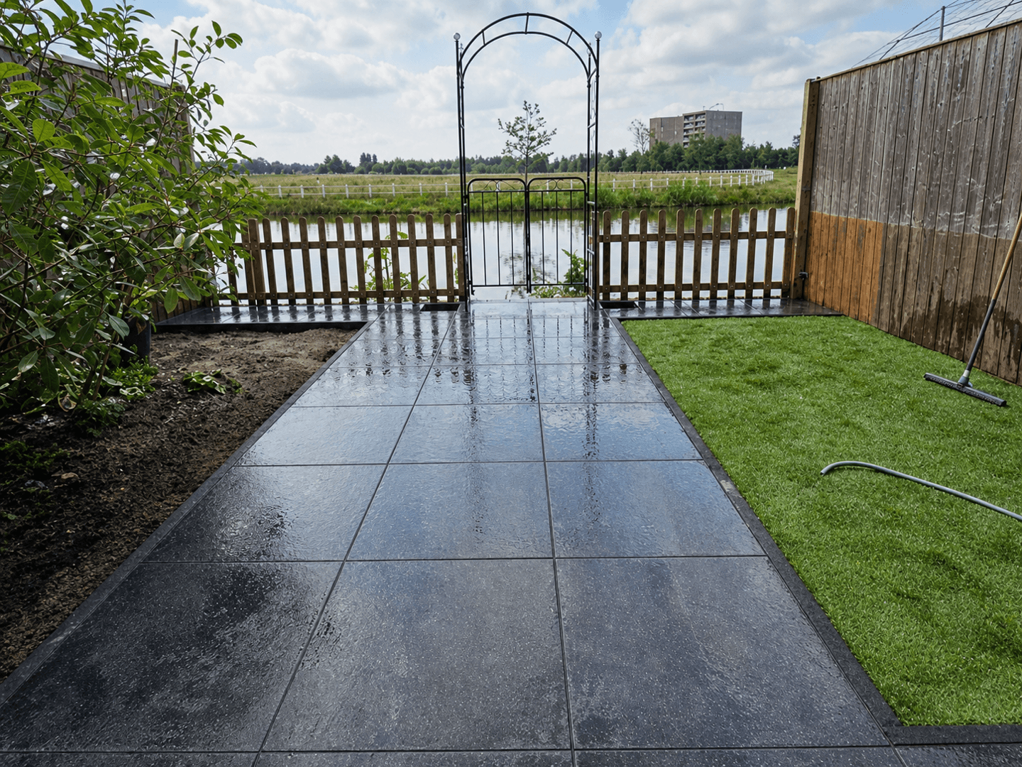 Nieuw aangelegd terras aan het water, met grote keramische tegels, kunstgras en een sierlijke tuinboog.