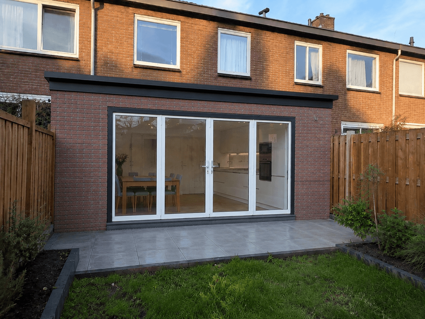 Afgewerkte keukenaanbouw aan een bakstenen rijwoning in Nederland, met tuindeuren, grijs porselein terras en een klein gazon.