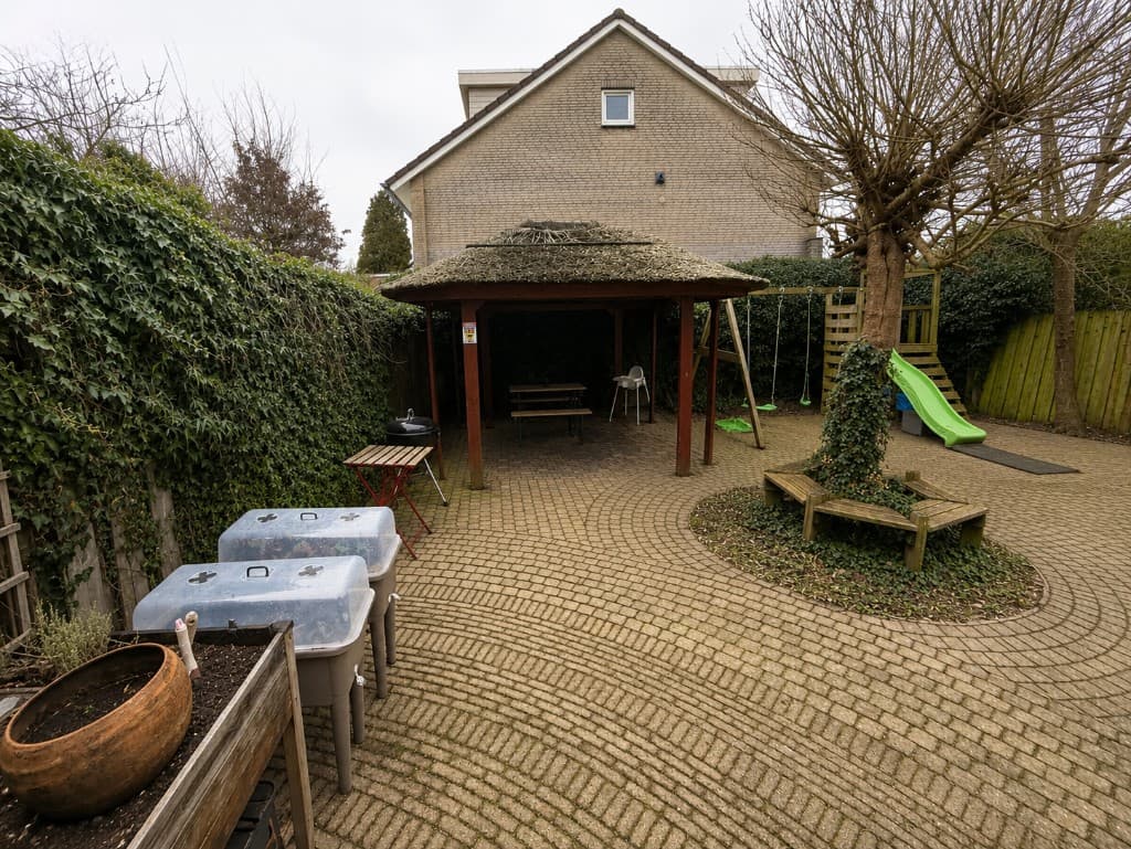 Originele achtertuin vóór de verbouwing, met gebakken tegels, houten gazebo en speeltoestel.