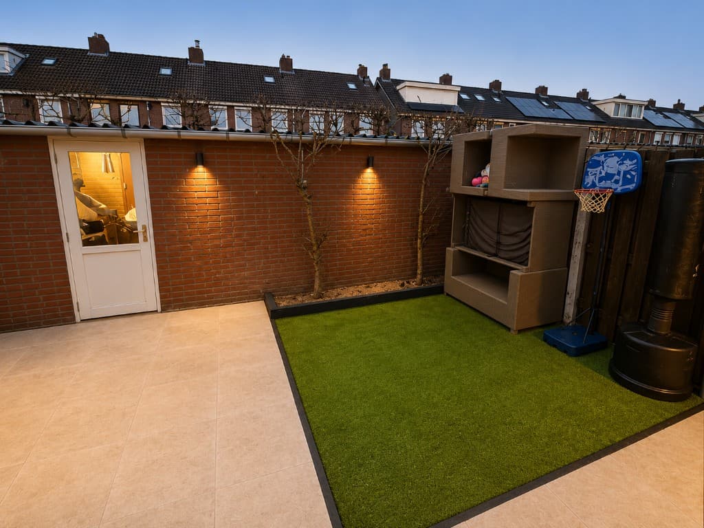 Renovated garden and patio at a home in the Eindhoven area, with paving, artificial grass and outdoor lighting at dusk.