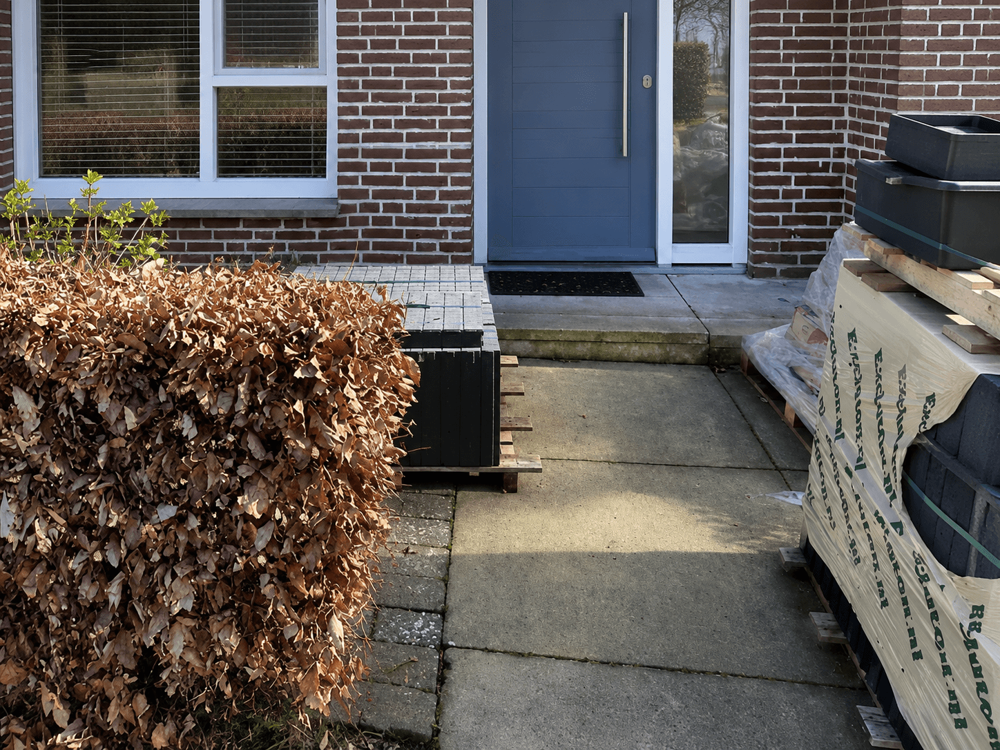 Voortuin vóór de verbouwing, met donkere tegels, een uitgebloeide haag en opgeslagen materiaal.