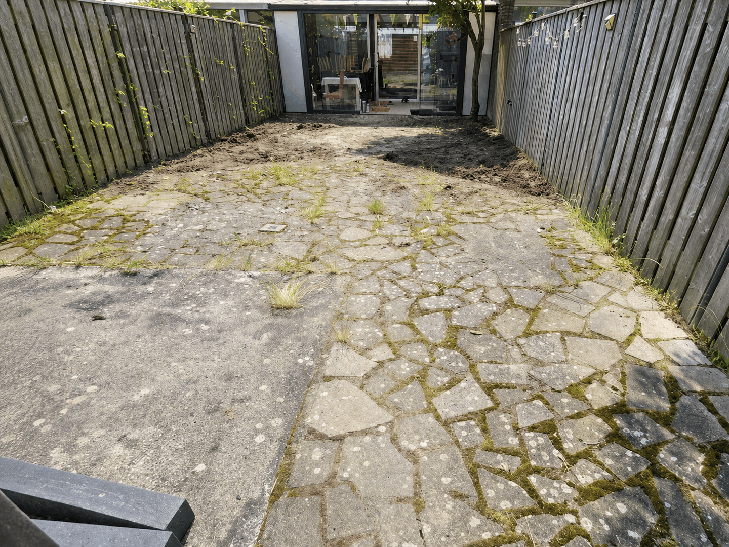 Smalle stadstuin vóór de renovatie, met kapotte flagstones vol mos en onkruid.