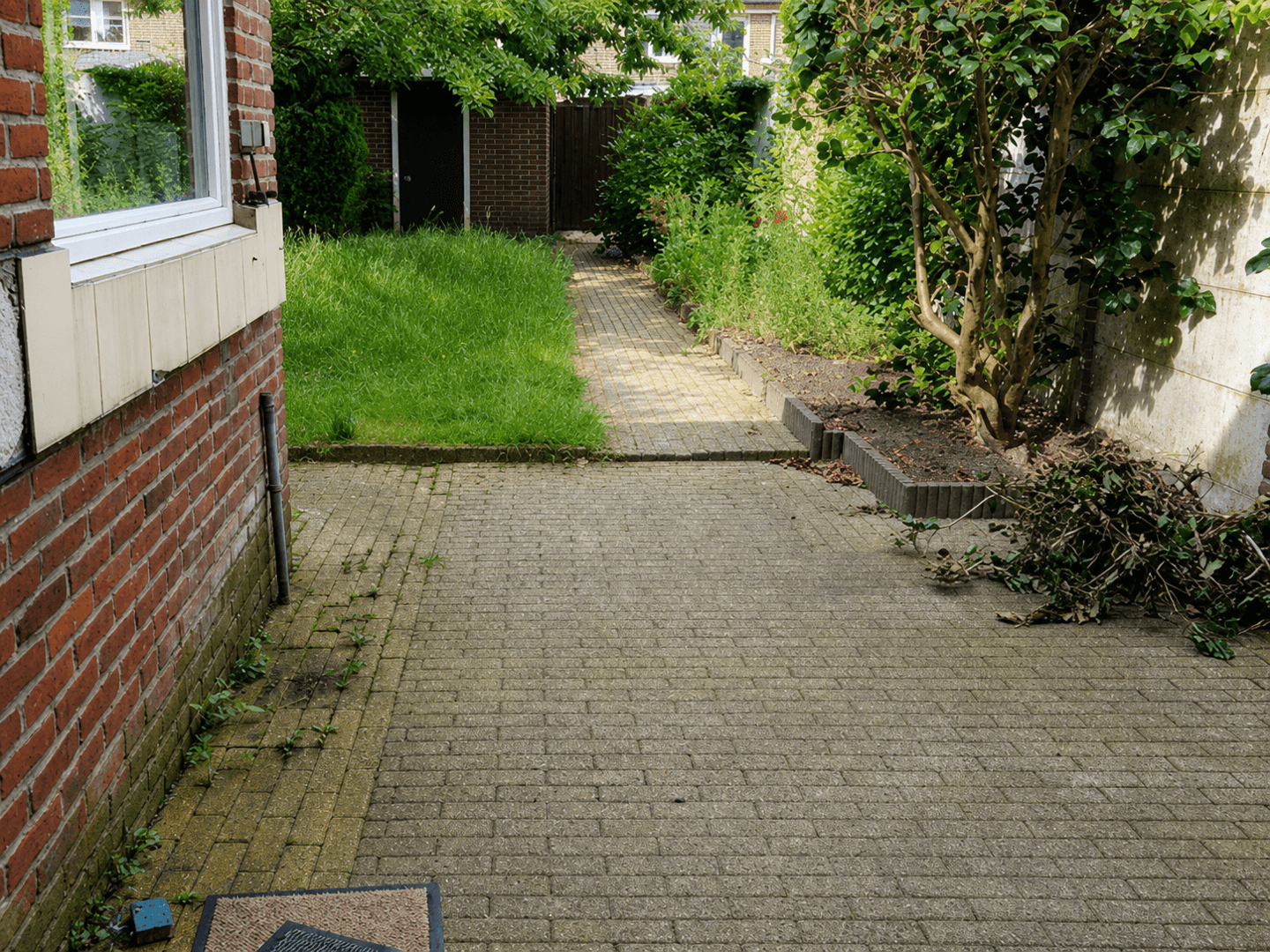 Achtertuin vóór de heraanleg, met begroeide gebakken klinkers en achterstallig groen.